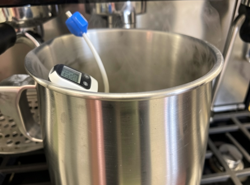 A probe thermometer clipped to the side of a stainless milk pitcher reading just below 60 degrees Celsius while steaming
